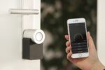 Ein schlankes, weißes Nuki Keypad mit beleuchtetem Zahlenfeld ist neben einer modernen, smarten Haustür montiert. Eine Hand tippt eine PIN ein, während die Tür leicht geöffnet ist – ein Symbol für komfortablen, schlüssellosen Zutritt im smarten Zuhause.