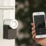 Ein schlankes, weißes Nuki Keypad mit beleuchtetem Zahlenfeld ist neben einer modernen, smarten Haustür montiert. Eine Hand tippt eine PIN ein, während die Tür leicht geöffnet ist – ein Symbol für komfortablen, schlüssellosen Zutritt im smarten Zuhause.